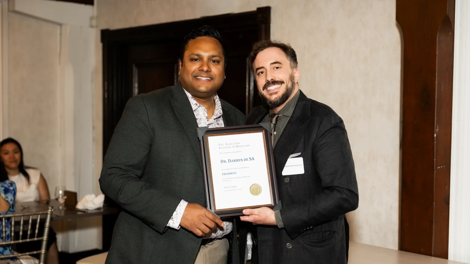 A framed certificate recognizing Dr. Darren de SA as HAM President, held up by two physicians at the Annual General Meeting